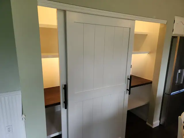 Custom pantry with sliding white barn door open to lit shelves and stained wood countertop
