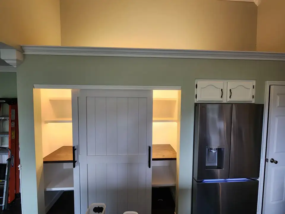 Home remodeling and repair by McMichael Home Repair: walk-in kitchen pantry with a white sliding barn door, lit white shelves and stained wood counters beside a stainless refrigerator
